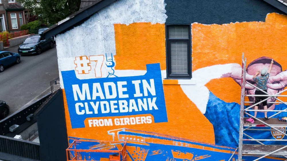 A painter working on a large orange and blue mural of John McGinn on the side of a house