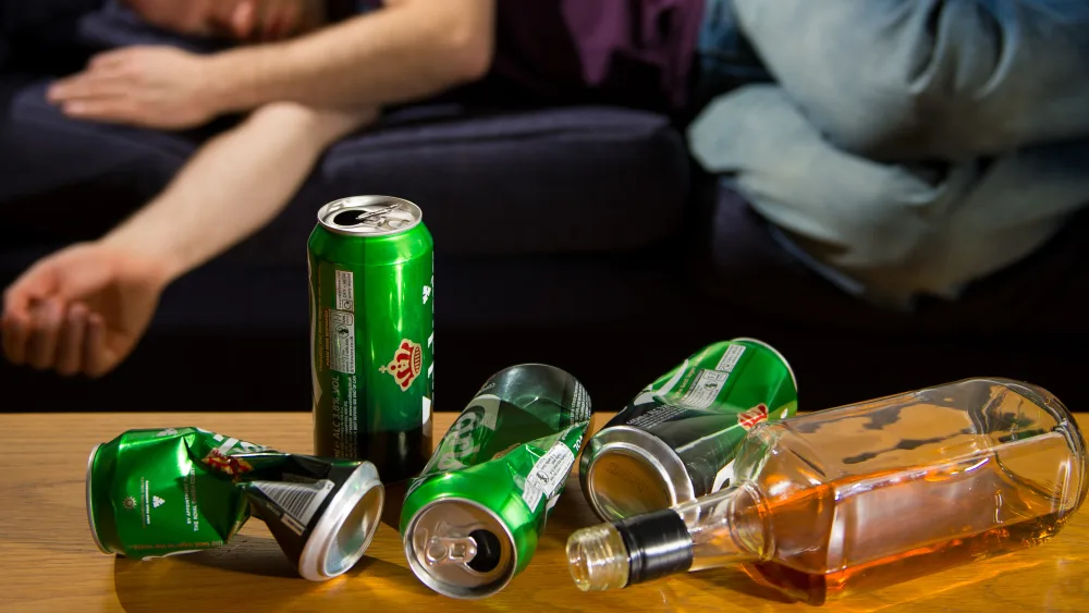 A man lies on a sofa with a table of empty alcohol bottles and cans in the foreground