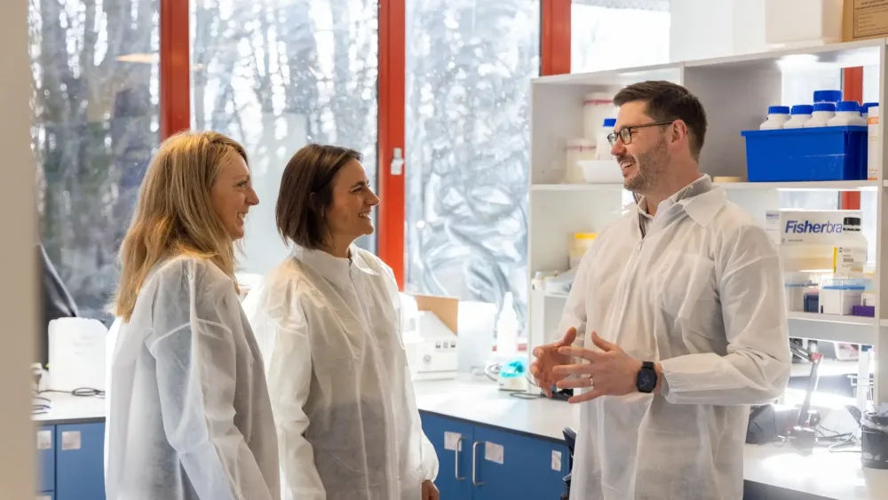 Jane Reoch and Ailsa Young standing opposite Douglas Martin and chatting to him, with all three wearing while lab coats