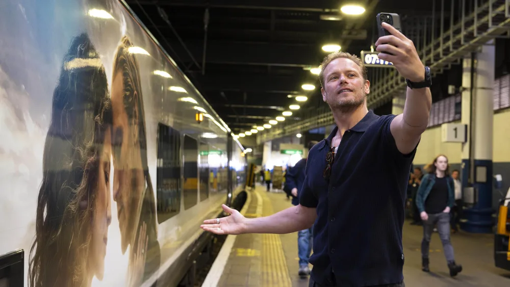 Sam Heughan takes a selfie of himself in front of a train featuring an Outlander-themed wrap