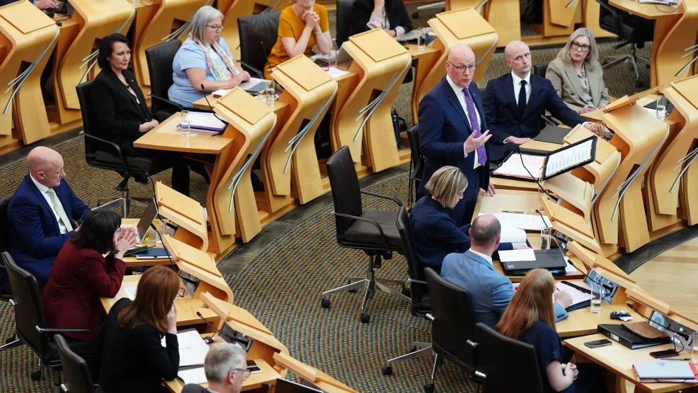 John Swinney standing while speaking in Holyrood, with a number of MSPs seated at their desks