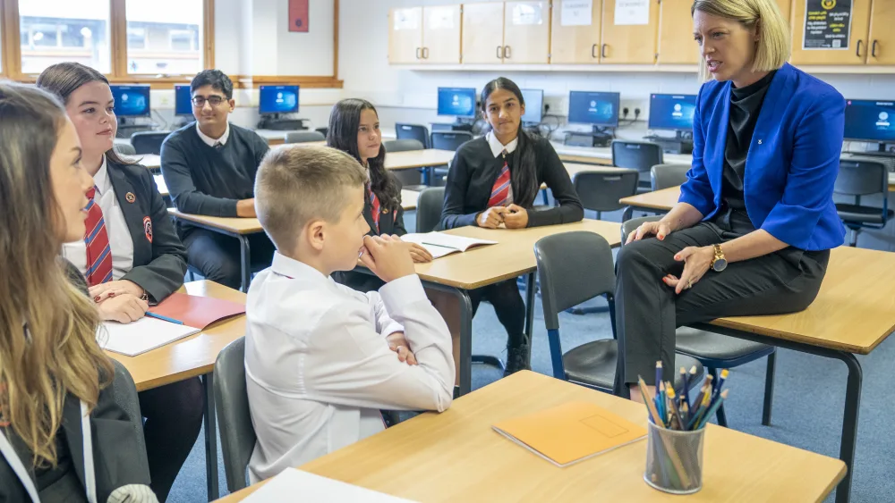 Jenny Gilruth sitting on a desk in a classroom while speaking to a group of senior school pupils
