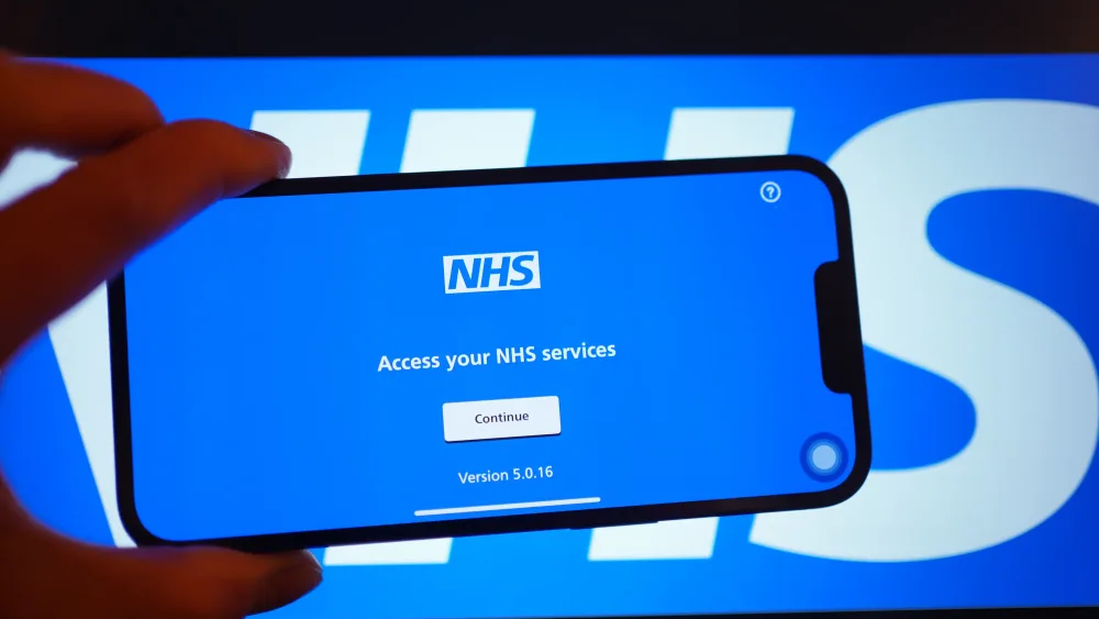 NHS app displayed on a smartphone in front of a large NHS sign