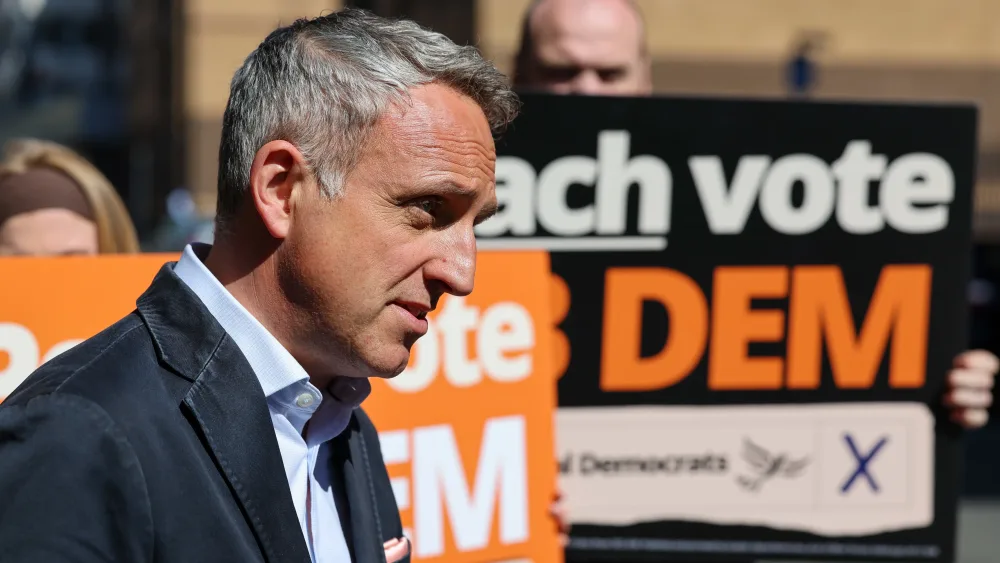 Side view of Alex Cole-Hamilton speaking outside, in front of a group of Lib Dem activists waving placards