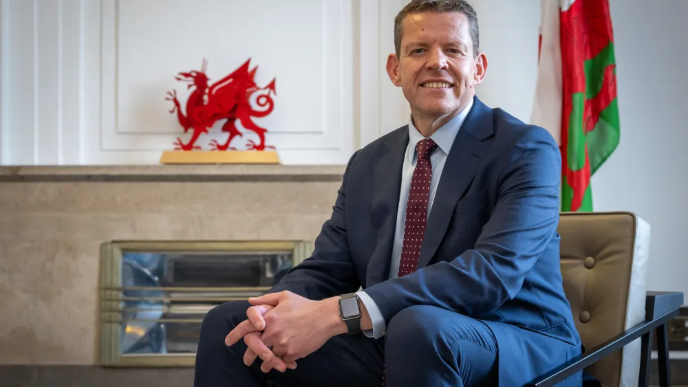 Rhun ap Iorwerth smiling while seated in an armchair, the Welsh flag behind him