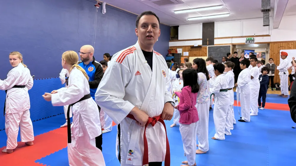 Anas Sarwar wearing a white karate gi with a red belt while young people behind him take part in a karate session