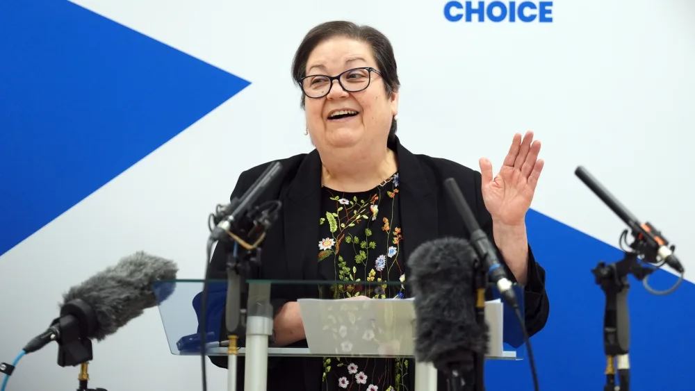 Dame Jackie Baillie speaking from a lectern in front of a large Scottish flag
