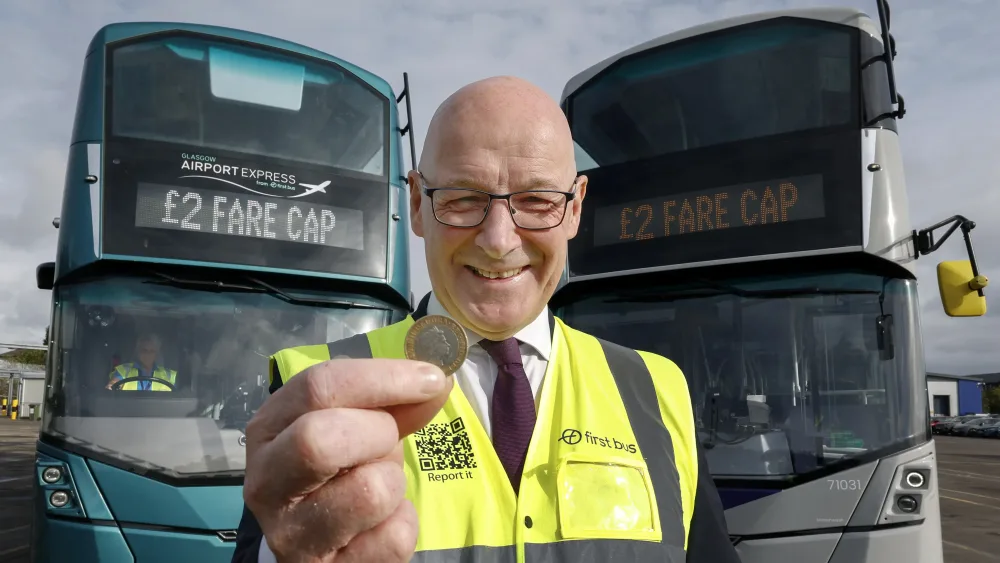 John Swinney holds a two-pound coin while posing in front of two buses