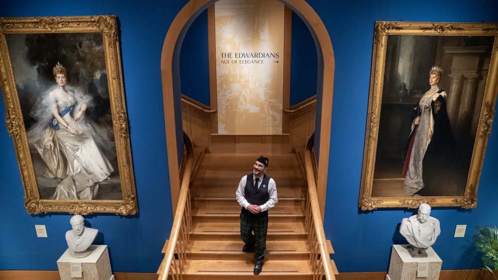 A man walking down a staircase, flanked by large paintings of Queen Alexandra and Queen Mary
