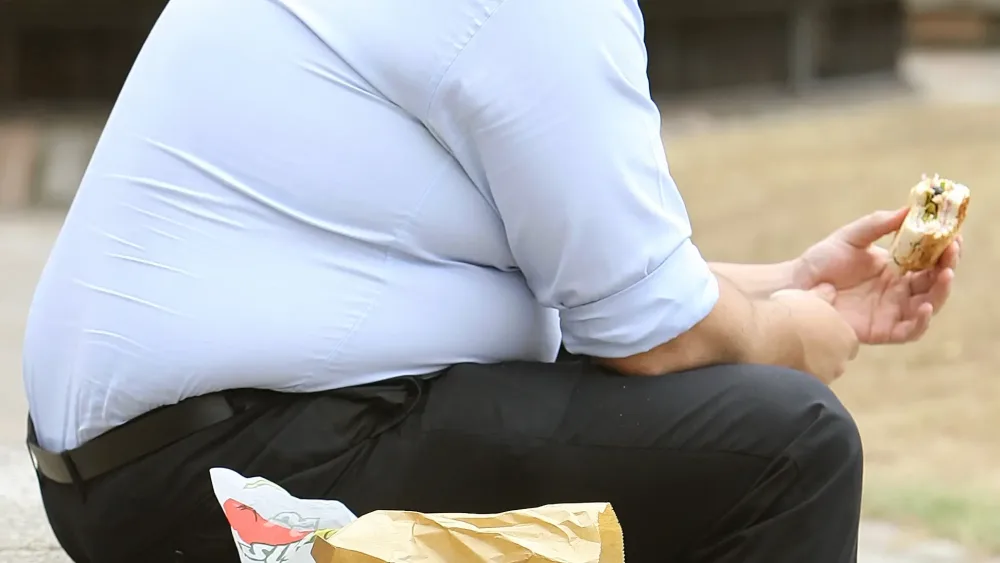 An overweight man sitting on a bench eating a burger