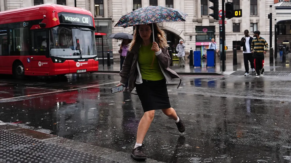 A woman steps over a puddle as she crosses a road in the rain, with a red London bus in the background